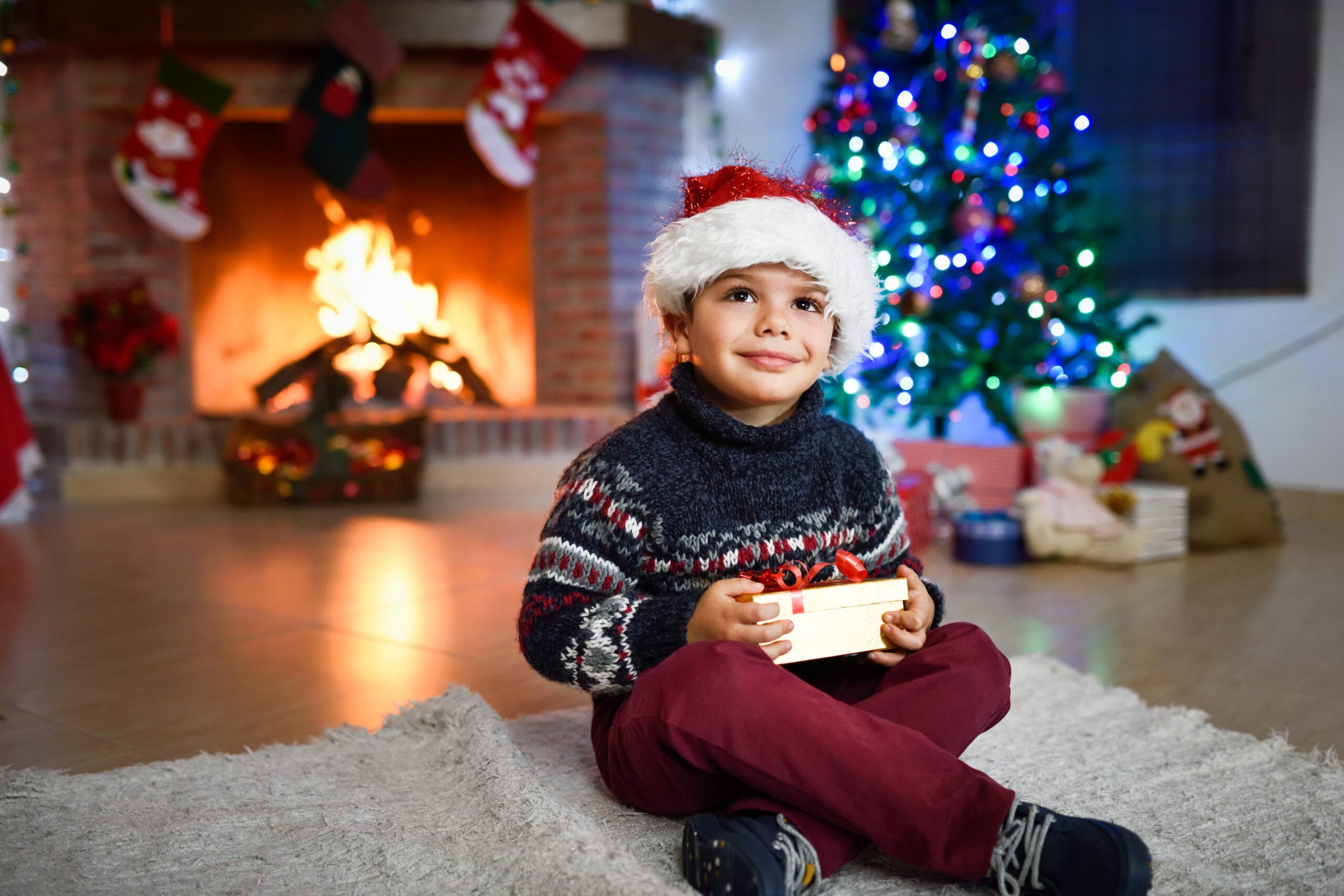 Happy little boy in a room decorated for Christmas with golden gift box wearing Santa hat