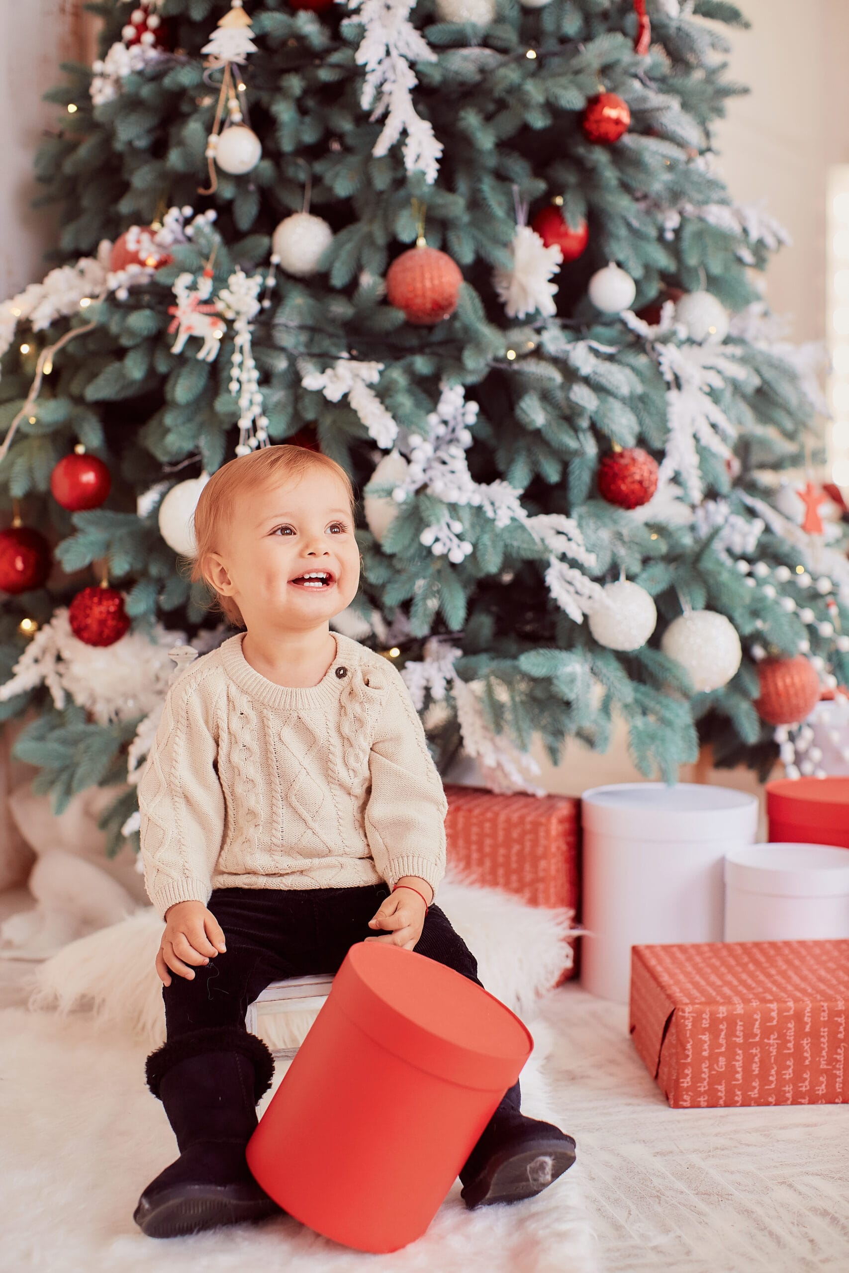 Winter holidays decorations. Warm colors. Beautiful little girl plays with present boxes before a rich decorated Christmas tree in a cozy beige room